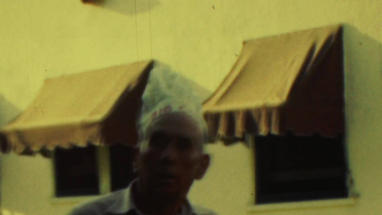 Chef-hatted man faces the camera from behind a brick grill and metal rail.
