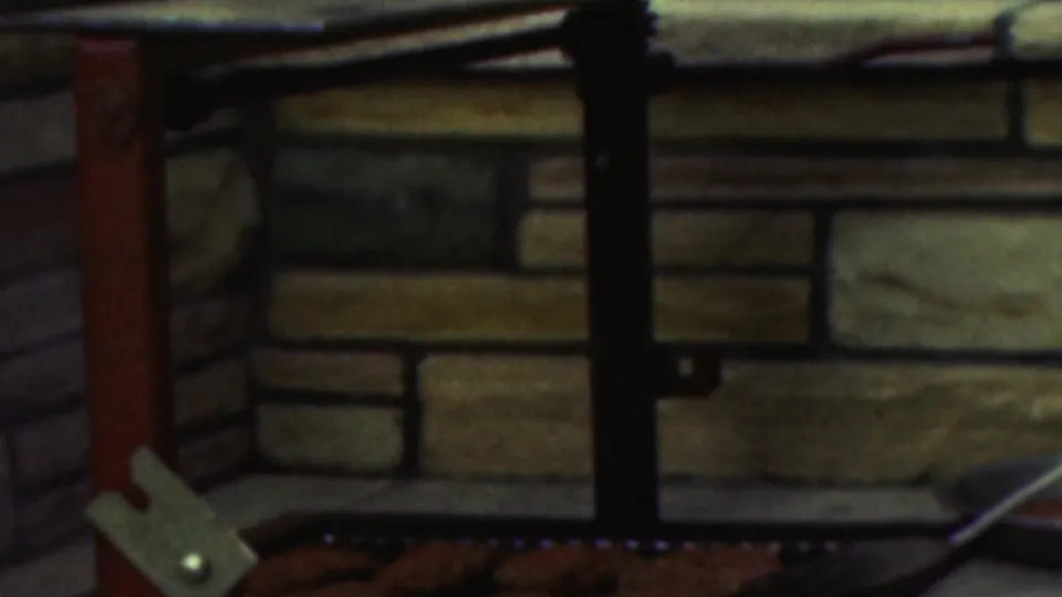 A closer view shows the brick grill structure, dark metal railings, cooking surface, food on the grill, and a metal spatula near the lower edge of the 8mm frame.
