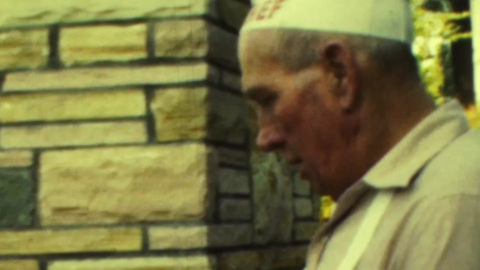 An older man in a white paper hat and light shirt stands in profile beside the stone outdoor fireplace in this 1949 Seaside, New Jersey home-movie frame.