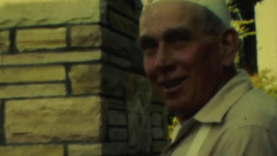 The older man turns toward the camera and smiles while standing near the stone backyard fireplace, with warm foliage visible behind him.