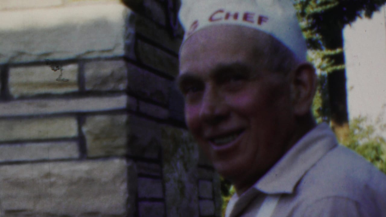 In 1949 Seaside, New Jersey home-movie footage, a smiling man wearing a white chef hat with red lettering stands near a stone wall outdoors.