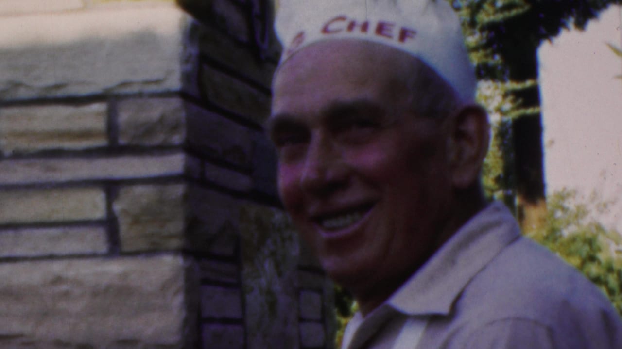 Man in a white chef hat smiles near stonework and greenery.
