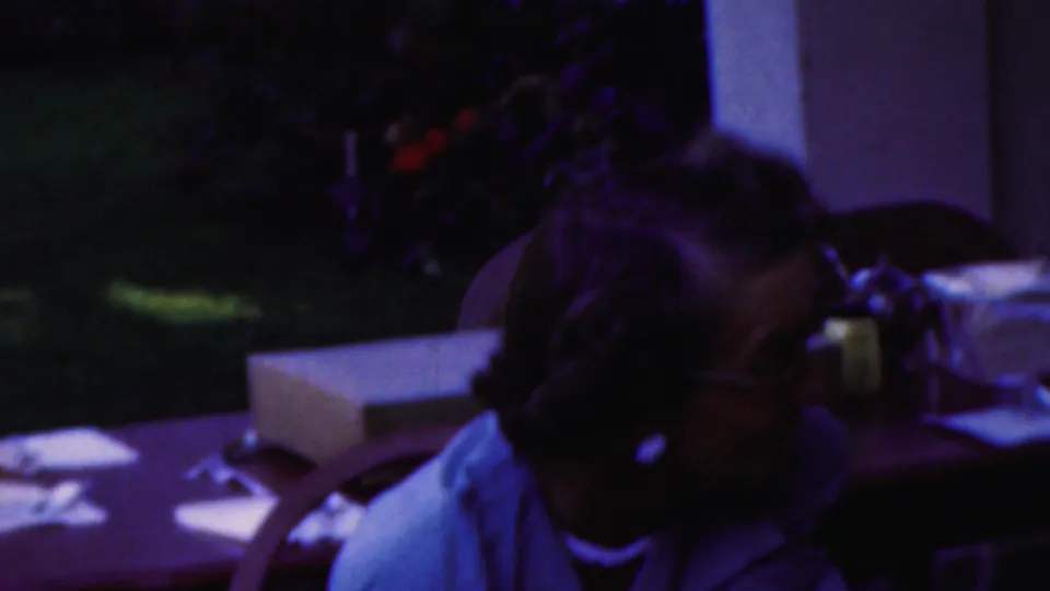 A woman with dark hair sits at an outdoor table in the 1949 Seaside, New Jersey home-movie footage, with papers or napkins and patio furniture nearby.