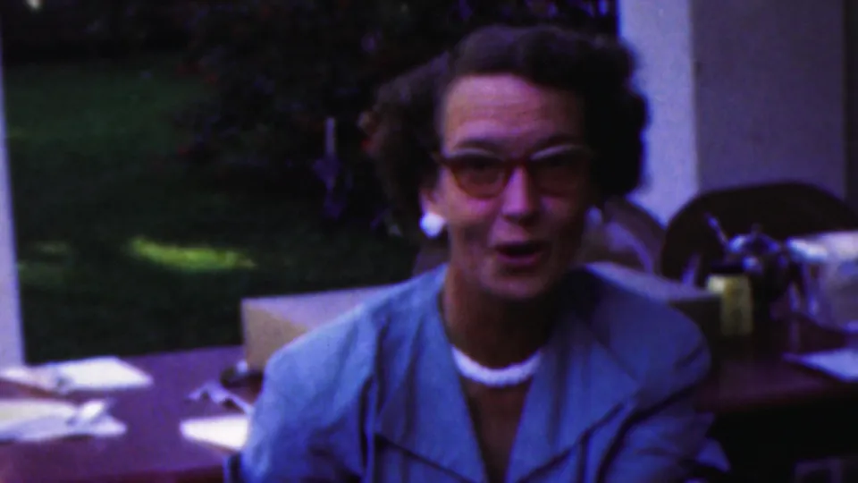 A woman in glasses faces the camera from the party table, with the soft color and grain of 1949 8mm home-movie footage.