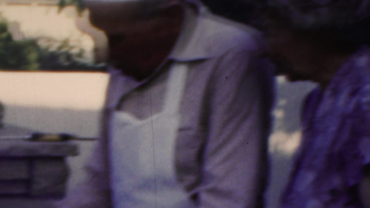 In 1949 Seaside, New Jersey home-movie footage, a man in a white apron stands near an outdoor cooking area while a woman in a patterned dress is partly visible beside him.