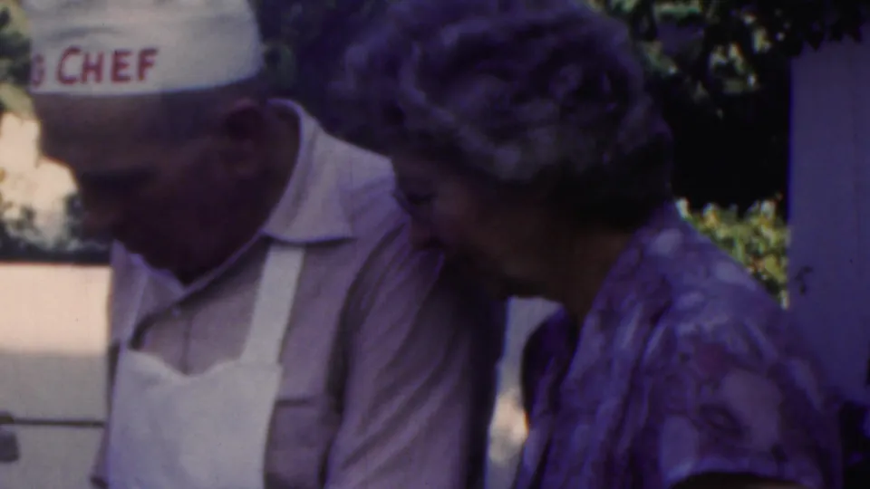 In the Seaside, New Jersey 1949 cookout scene, a man wearing a “BIG CHEF” hat and white apron leans near a woman in a patterned dress.