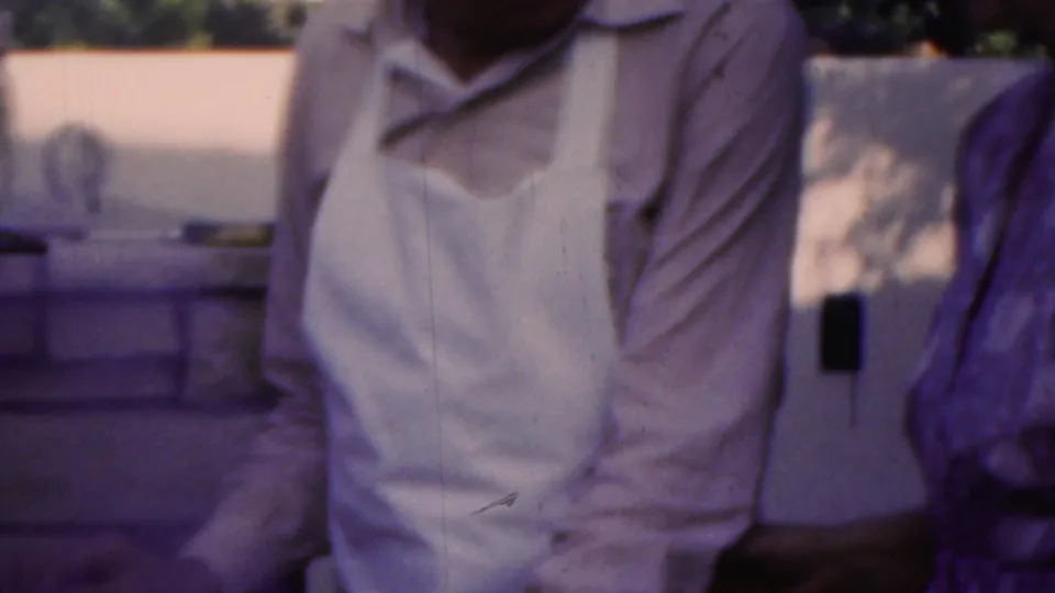 In Seaside, New Jersey home-movie footage from 1949, an older man in a light shirt and white apron stands beside an older woman in a patterned dress on a porch.