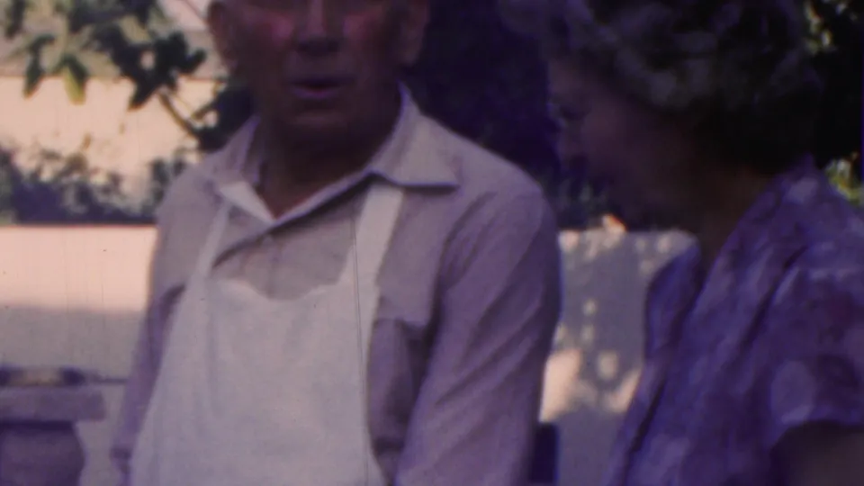 An older man in a white apron appears to speak with an older woman beside a porch, with trees and daylight behind them in the 1949 Seaside, New Jersey home-movie scene.