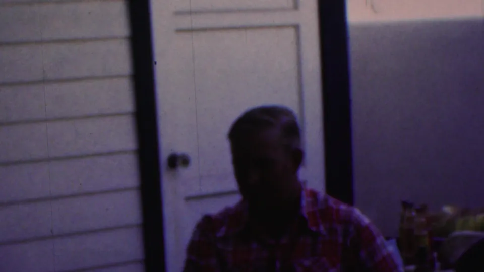 A darkly exposed 1949 Seaside, New Jersey frame shows an older person near a doorway and exterior siding, with porch objects partly visible in the background.