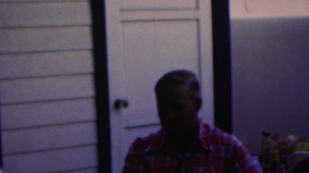 In this 1949 Seaside, New Jersey home-movie frame, a man in a plaid shirt sits at a table in low light against a pale paneled wall and door.