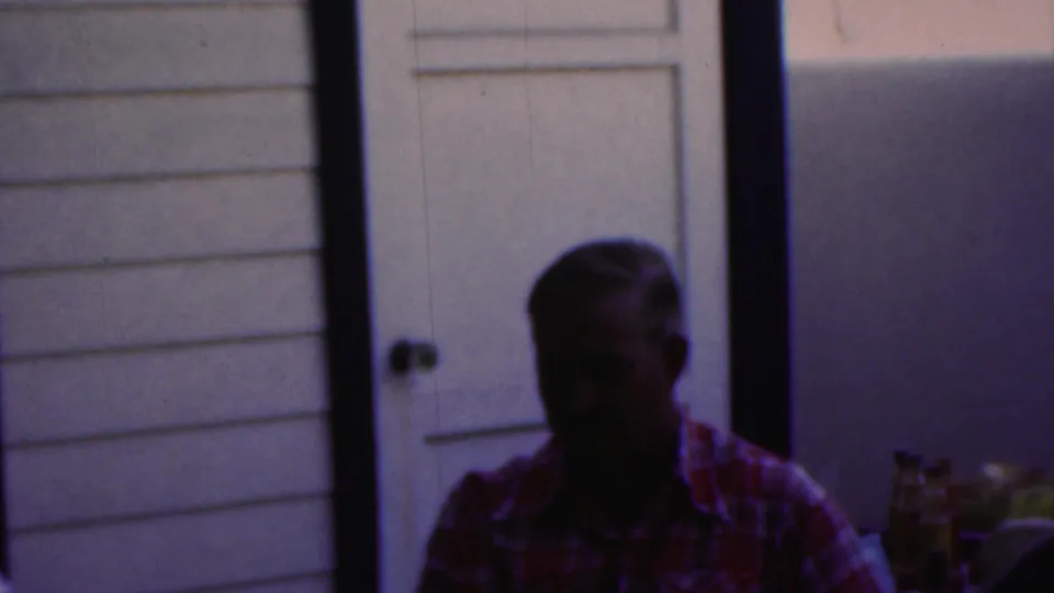 In this 1949 Seaside, New Jersey home-movie frame, a man in a plaid shirt sits at a table in low light against a pale paneled wall and door.