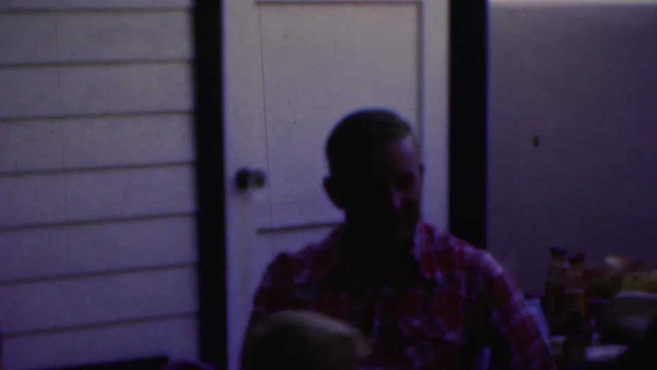 A seated man in a red plaid shirt appears beside a table with bottles or condiments, with a white door and paneled wall in the background.