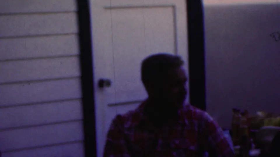 The man remains seated at the table and turns slightly, with the dim purplish 8mm exposure obscuring facial detail against the paneled wall.