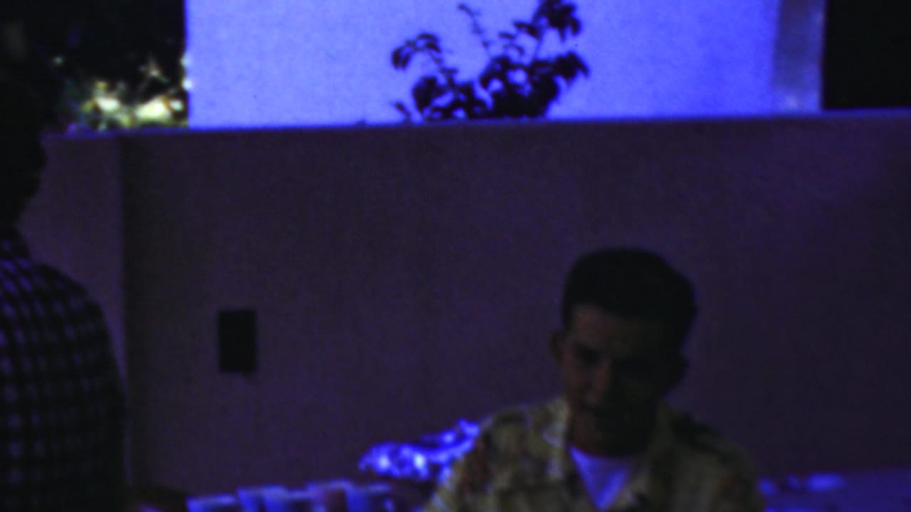 In Seaside, New Jersey home-movie footage from 1949, a young man in a patterned shirt sits at a table under strong blue-purple light with cups nearby.