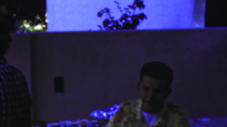 In Seaside, New Jersey home-movie footage from 1949, a young man in a patterned shirt sits at a table under strong blue-purple light with cups nearby.