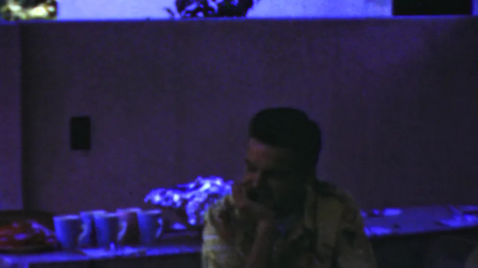 A young man in a patterned shirt turns to the side beside several cups on a table, with blue-purple light and a plain wall behind him in the Seaside, New Jersey footage.