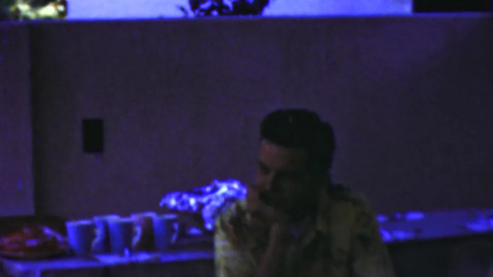 The young man sits with his hand near his face beside a row of cups, framed by a table surface, wall fixtures, and heavy blue-purple lighting in the 1949 Seaside, New Jersey clip.