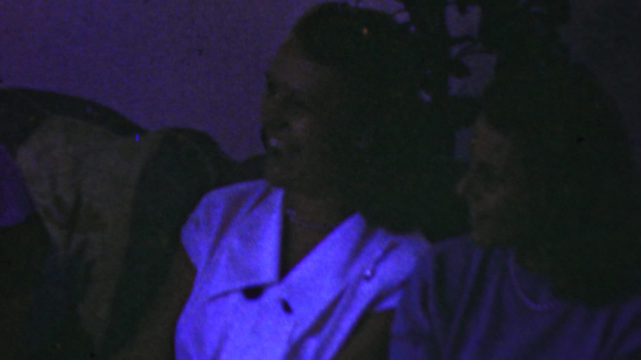 Several adults lean together on a couch in a dark room, with a woman in a white blouse at right.