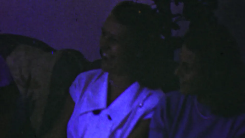 In the supplied 1949 Seaside, New Jersey setting, a small group of adults sits tightly together in a dark living room, including a woman in a white collared blouse turned toward another person.