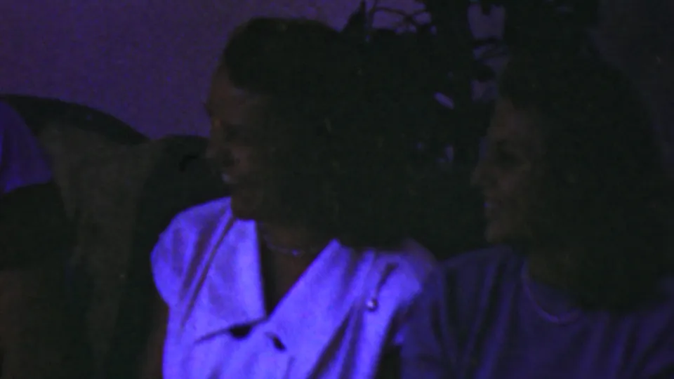 In the supplied 1949 Seaside, New Jersey home-movie scene, a woman in a white collared blouse turns toward seated companions in a low-lit living room with heavy blue-purple film color.