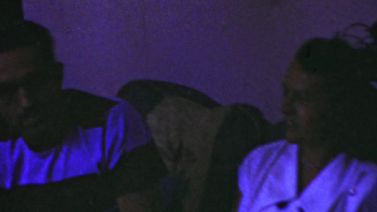 In metadata-supported 1949 Seaside, New Jersey home-movie footage, two adults sit close together in a dark interior under strong purple-blue lighting.