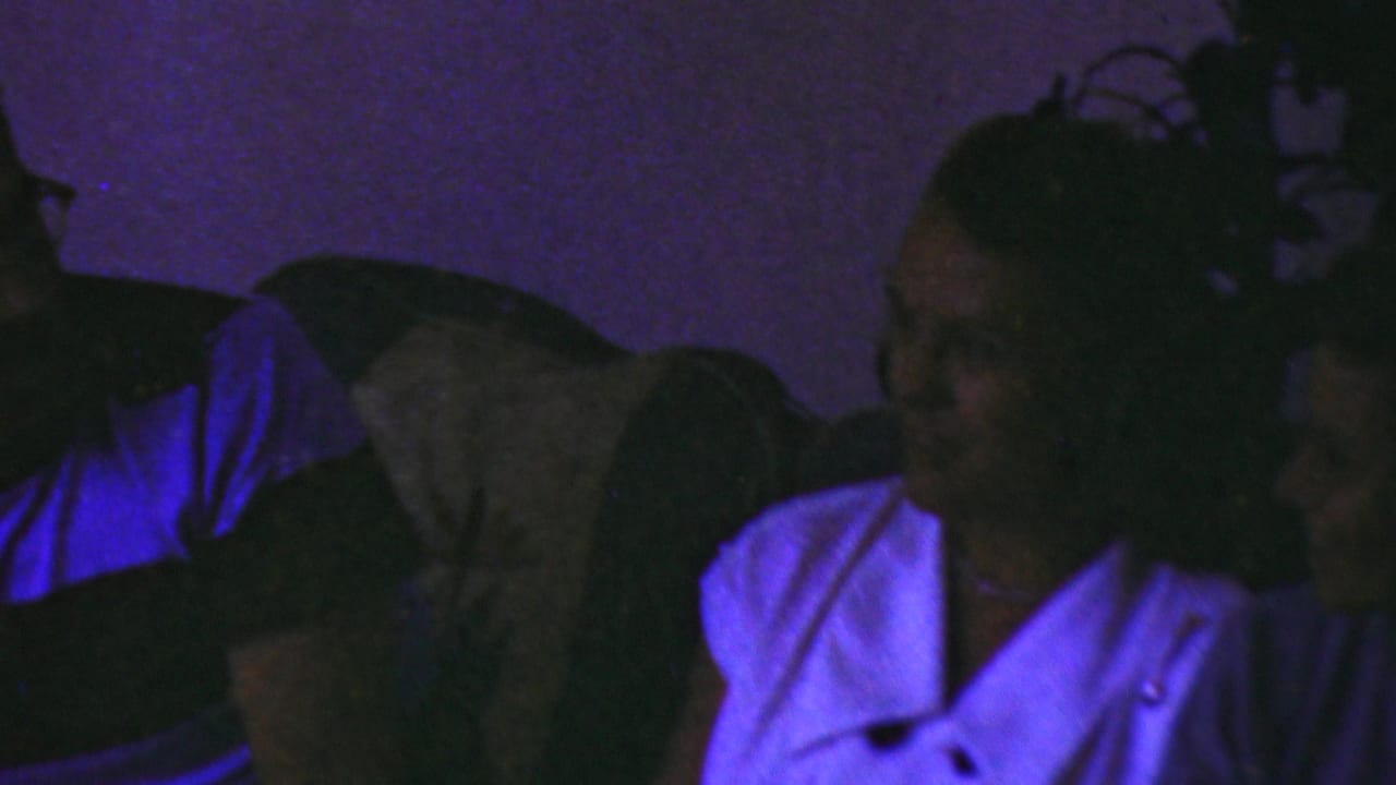 A woman in a light-colored top sits in a dim private interior in the Seaside, New Jersey home movie, her face partly obscured by low light and purple-blue color cast.