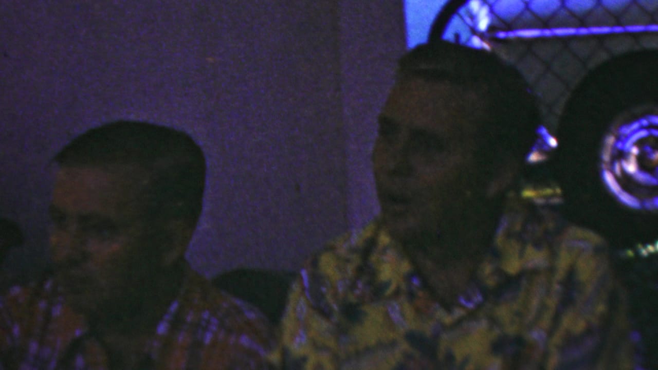 In 1949 Seaside, New Jersey home-movie footage, two adult men sit close together indoors, with a red plaid shirt at left and a yellow floral Hawaiian-style shirt at center under dim colored light.
