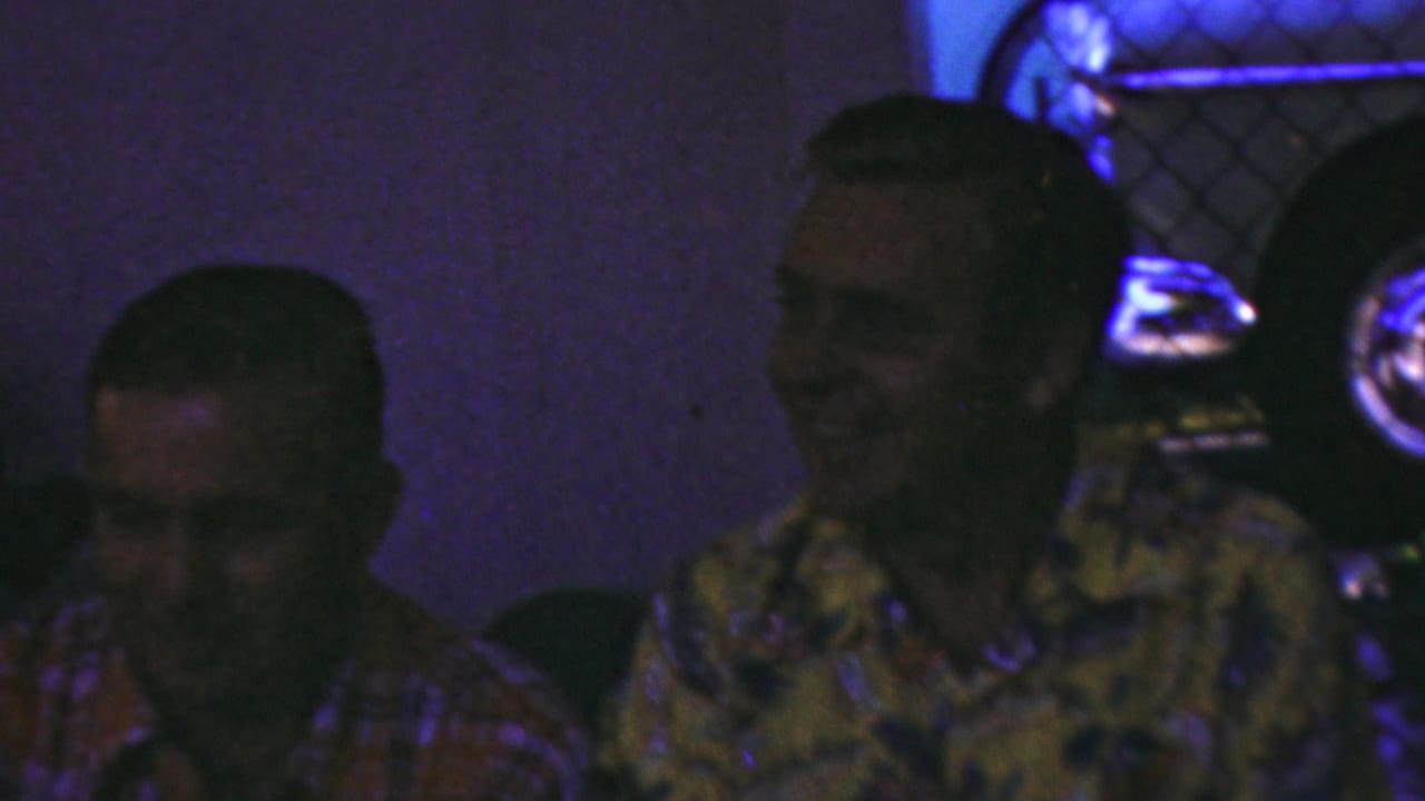 Three adult men seated indoors, with a smiling man in a yellow floral shirt beside a man in a red plaid shirt.