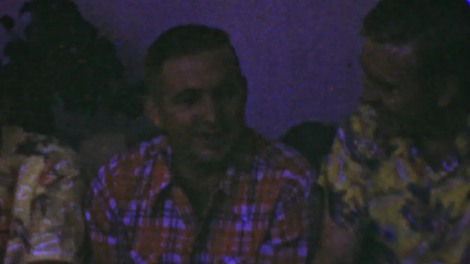 A wider 1949 Seaside, New Jersey home-movie frame shows several adult men seated close together in a dim room, including a man in a red plaid shirt facing camera and another in a yellow floral shirt turned sideways.