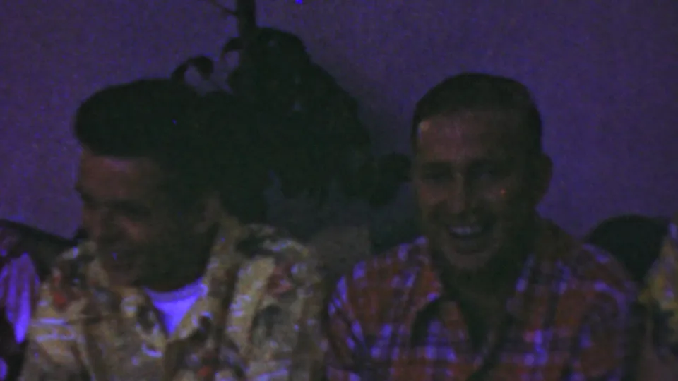 The final frame shows adult men seated together in the Seaside, New Jersey home movie, with a smiling man in a red plaid shirt at right and other men partially visible in patterned shirts under low purple-blue light.