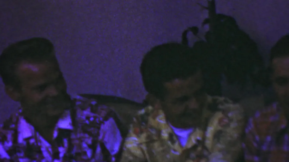 Three adult men appear to converse and smile in a dim indoor setting, with patterned shirts and dark vertical posts visible in the home-movie frame.