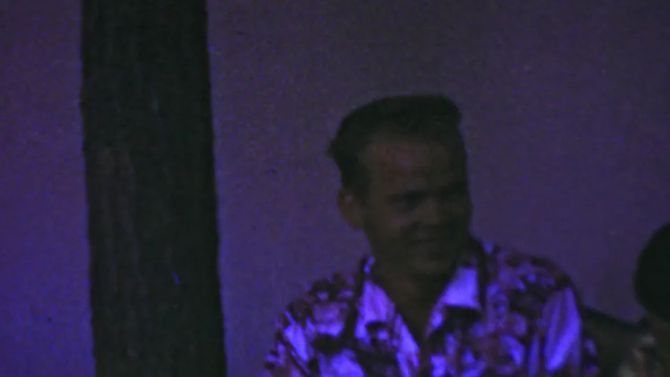 Two seated adult men are partly framed by dark vertical posts in a dim interior, with one face turned toward the camera and another figure lowered in shadow.