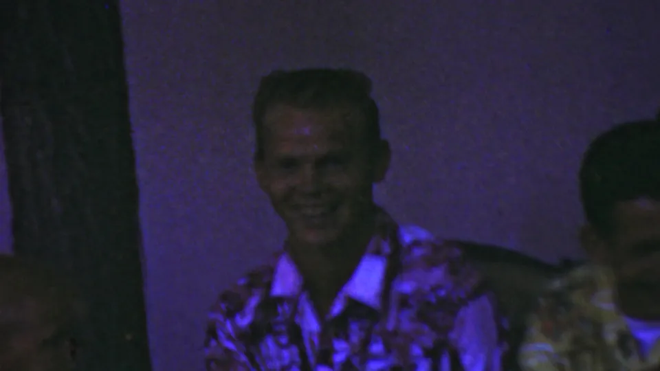 A smiling adult man in a patterned shirt sits in low purple interior light while another man is partly visible at the edge of the frame.