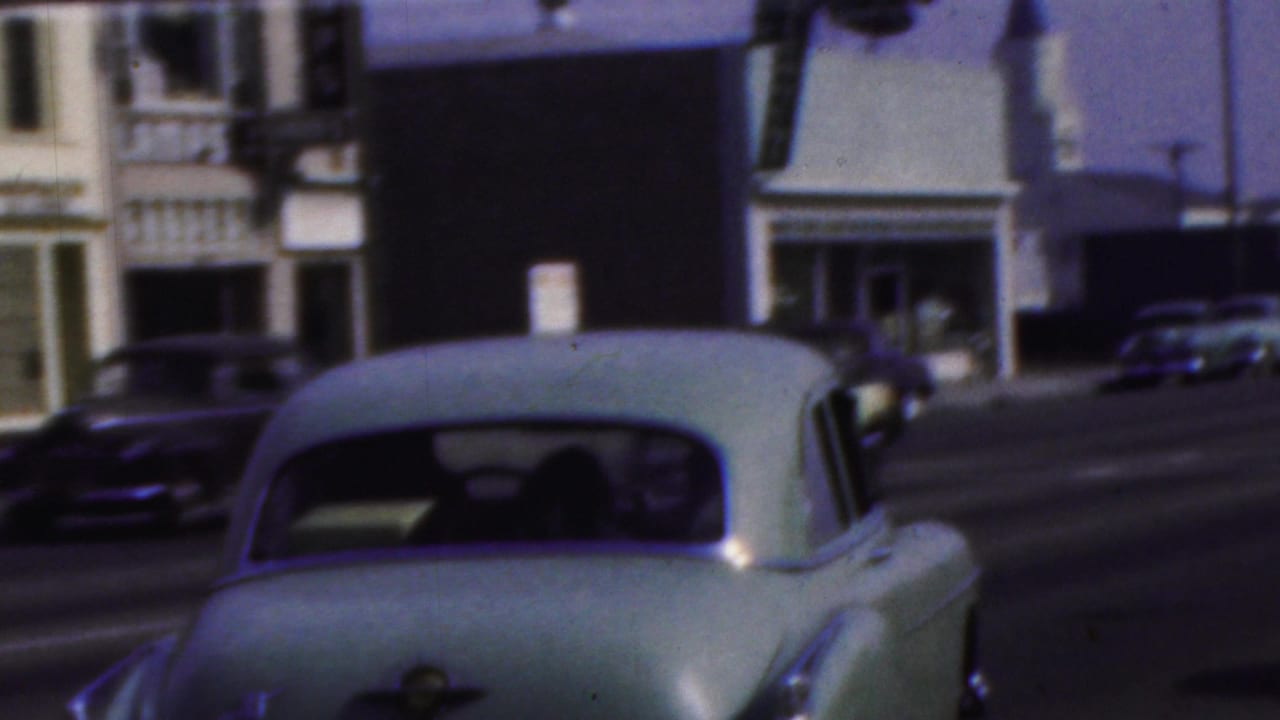 Rear view of a pale sedan passing storefronts with other cars nearby.