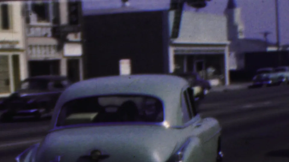 A pale sedan drives past storefronts and parked or passing cars in Seaside, New Jersey, as shown in the supplied 1949 8mm footage.