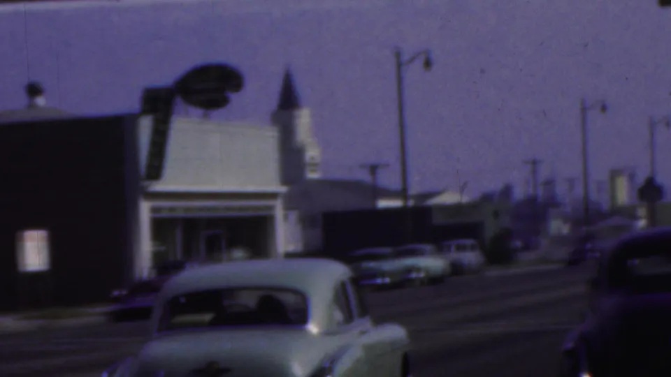 The pale sedan continues down the street past storefronts, streetlamps, parked cars, and a pointed roof detail in the supplied Seaside, New Jersey home-movie footage.