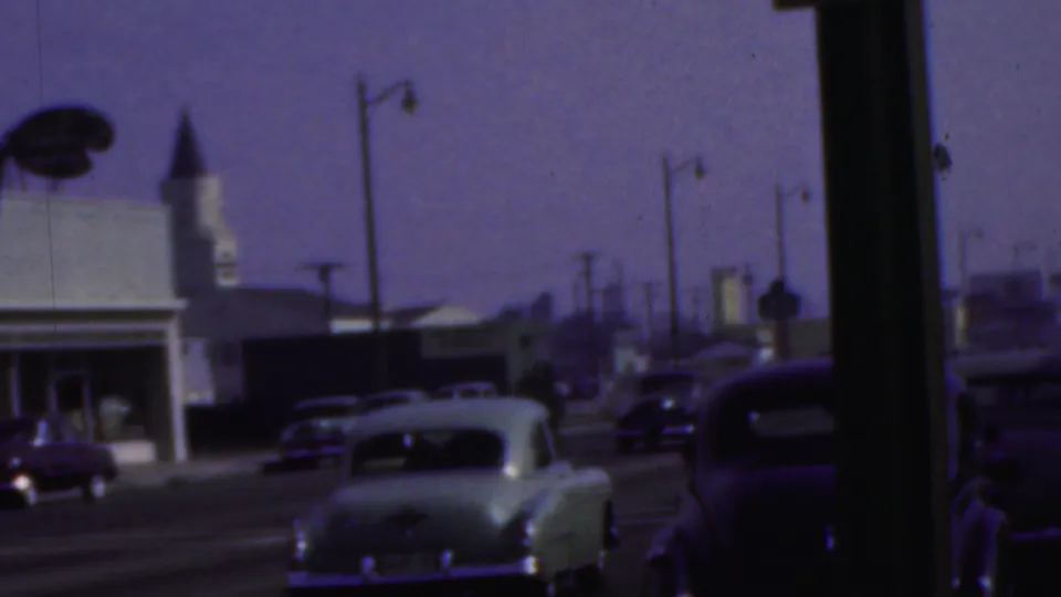 A wider 8mm frame shows the pale sedan farther down the Seaside, New Jersey street with parked cars, roadside buildings, streetlamps, and a visible sign form.