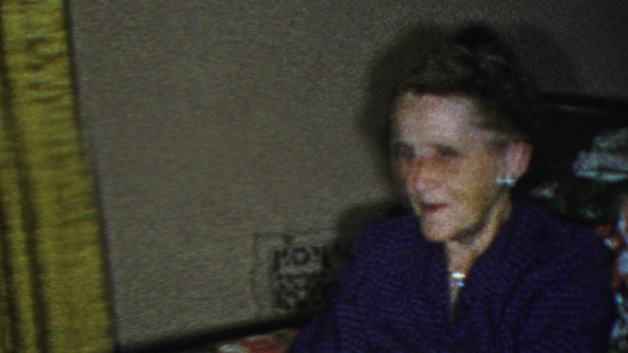 Elderly woman seated indoors beside green patterned curtains.