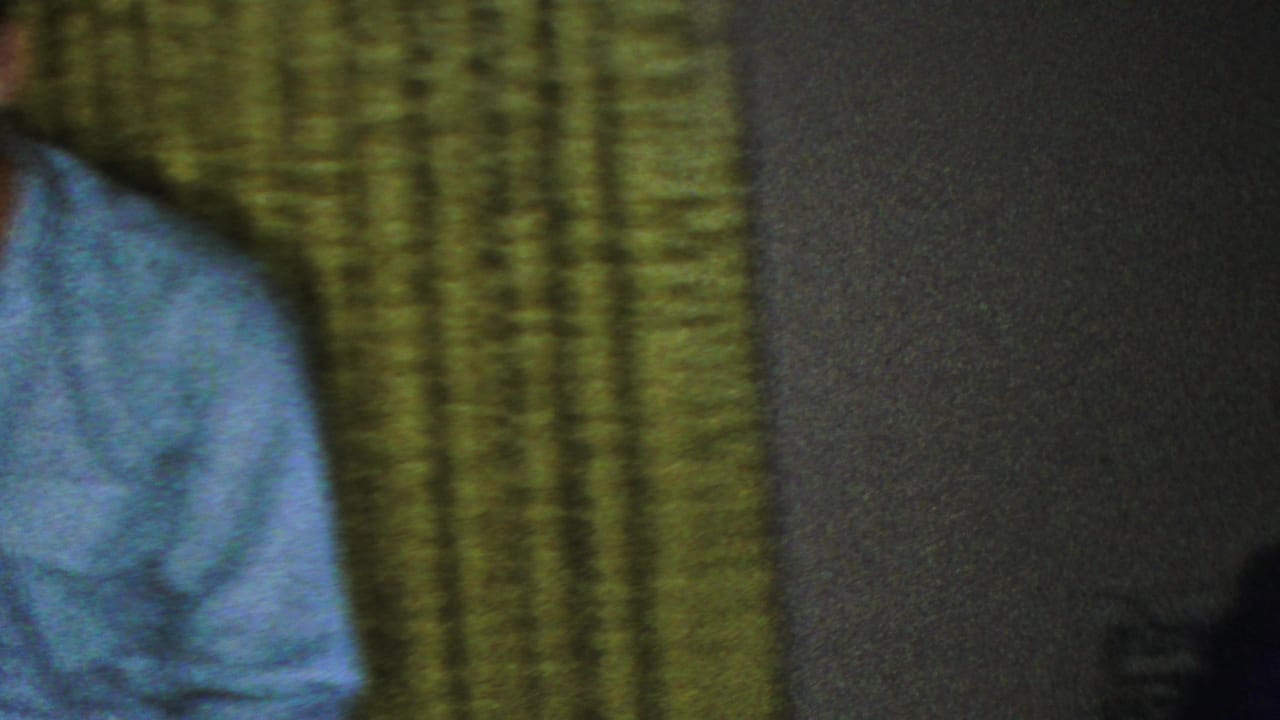 A man in a pale blue shirt leans or sits close to green patterned curtains in a dim private interior from the Seaside, New Jersey home-movie clip.