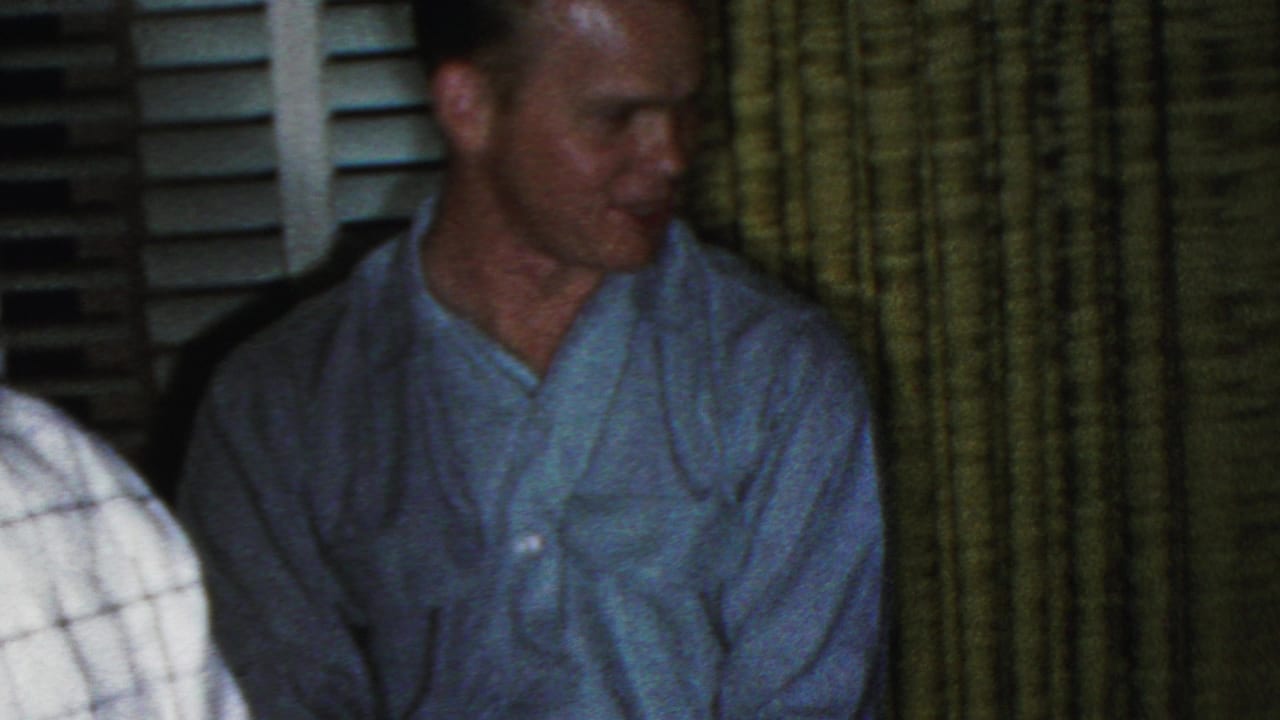 A seated man in a pale blue shirt faces toward the room beside green curtains and window shutters, with part of another person visible at the frame edge.