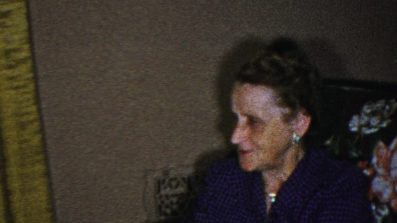 In this 1949 Seaside, New Jersey home-movie frame, an older woman wearing pearl earrings and a purple patterned dress sits on floral upholstery beside a green curtain.