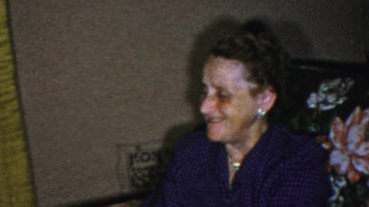 Older woman smiling while seated near floral upholstery in a dim interior.