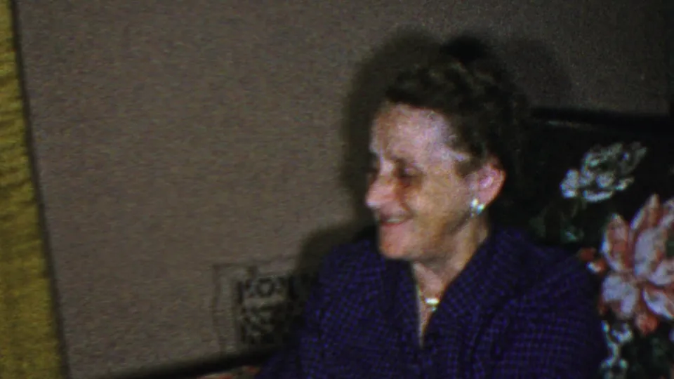 The older woman smiles in the living-room setting, with floral couch fabric, a dark chair back, and muted wall tones visible in the 1949 home-movie frame.