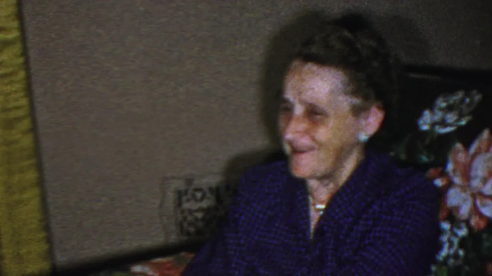 The seated older woman smiles on the floral couch while part of another woman appears at the lower right, reinforcing the candid family conversation described in the clip metadata.