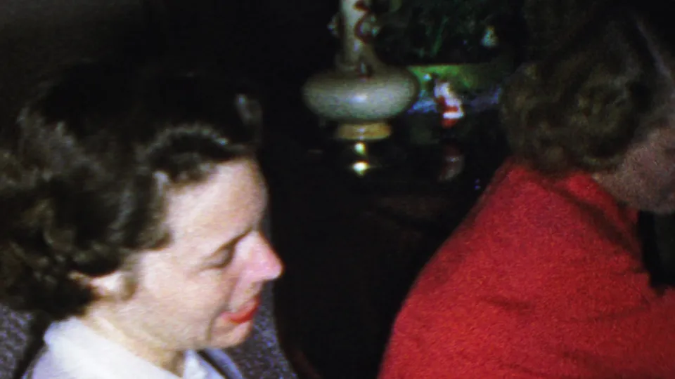 A wider 1949 Seaside, New Jersey home-movie frame shows the older woman partly obscured as two younger women, one in a light blouse and one in a red sweater, sit nearby with a lamp and floral couch visible.