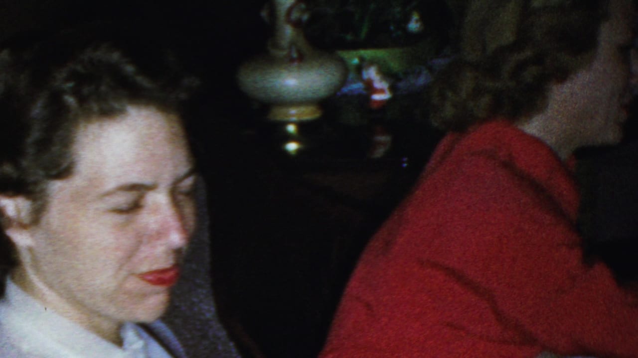 In a 1949 Seaside, New Jersey home-movie frame, a woman in a white collared blouse sits near a second woman in a red sweater, with a lamp and houseplants behind them.