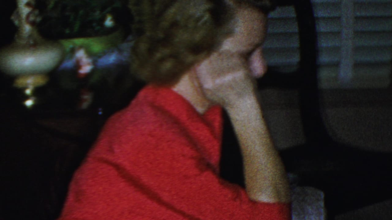 A woman in a red sweater rests her cheek against her hand in a dim private living room, with another seated figure and window shutters visible nearby.