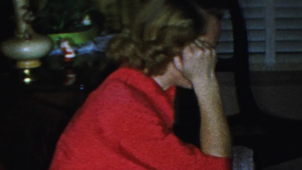 The woman in the red sweater appears in profile with her hand near her face, framed by indoor seating, a lamp, plants, and shuttered windows in the background.