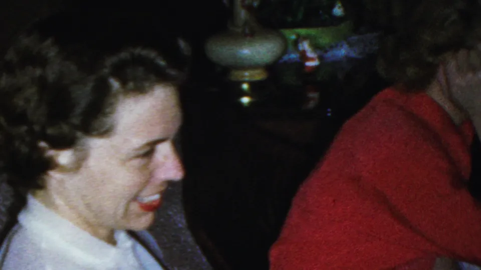 A smiling woman in a white collared blouse sits beside a woman in a red sweater in the 1949 Seaside, New Jersey living-room footage, with a lamp and plants behind them.