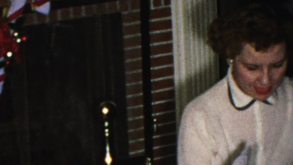The woman looks downward in front of a brick fireplace with a candy cane decoration, captured in warm, grainy 8mm home-movie color.